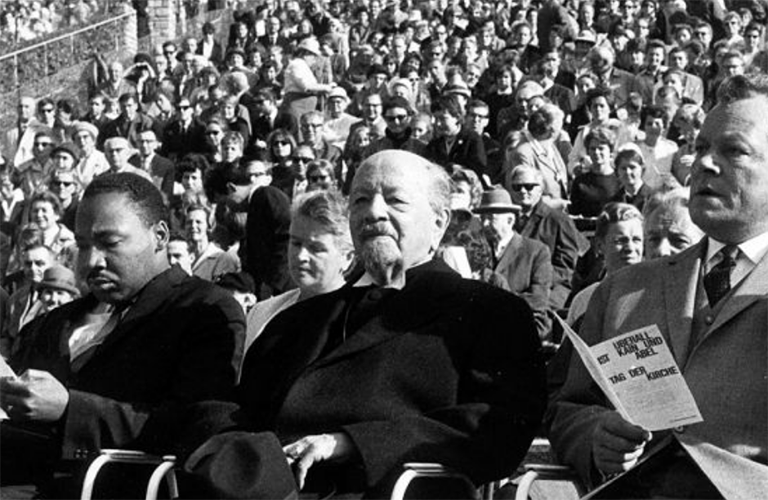 Black-and-white photo of Dr. Marin Luther King Jr. sitting at a rally in Berlin with German politicians