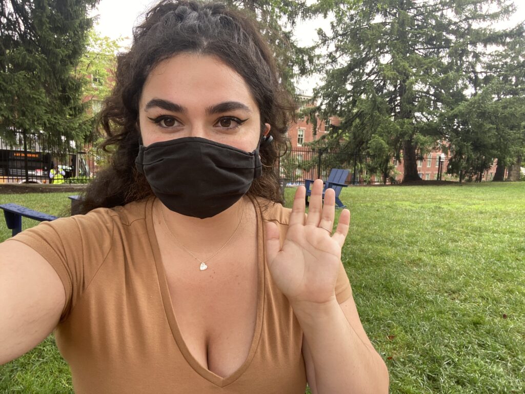 Alanna Cronk wearing a mask and waving to the camera while sitting on Healy lawn