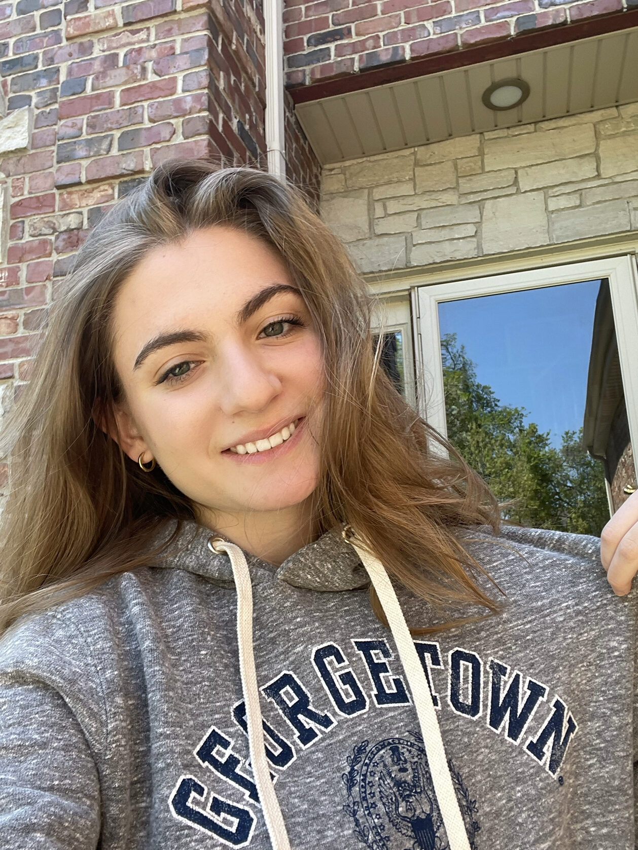 Ava smiles in her selfie as she pulls up one side of her grey hoodie so that you can see the Georgetown logo on it. 