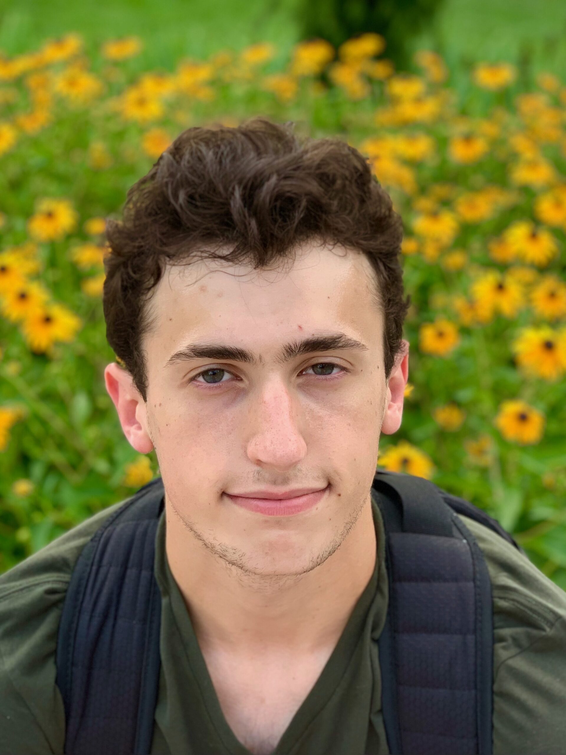 Epstein-Shuman looks into the camera as he sits in a field of yellow flowers. He is wearing a dark green shirt. 