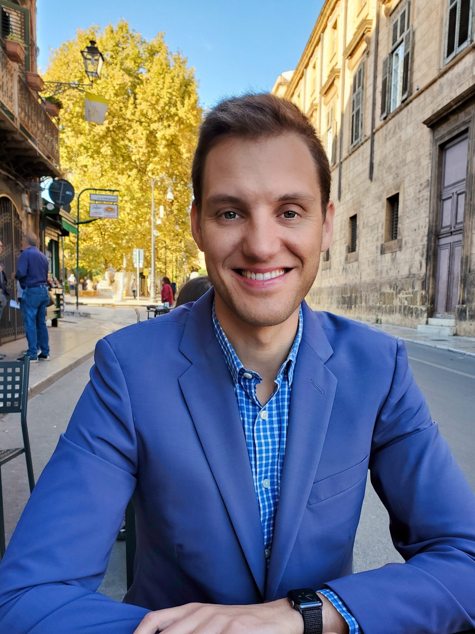 Kushlev smiling into the camera while he sits at a table outside. He is wearing a bright purpley-blue suit, the sky is bright blue, and there is a brilliantly yellow tree behind him. 