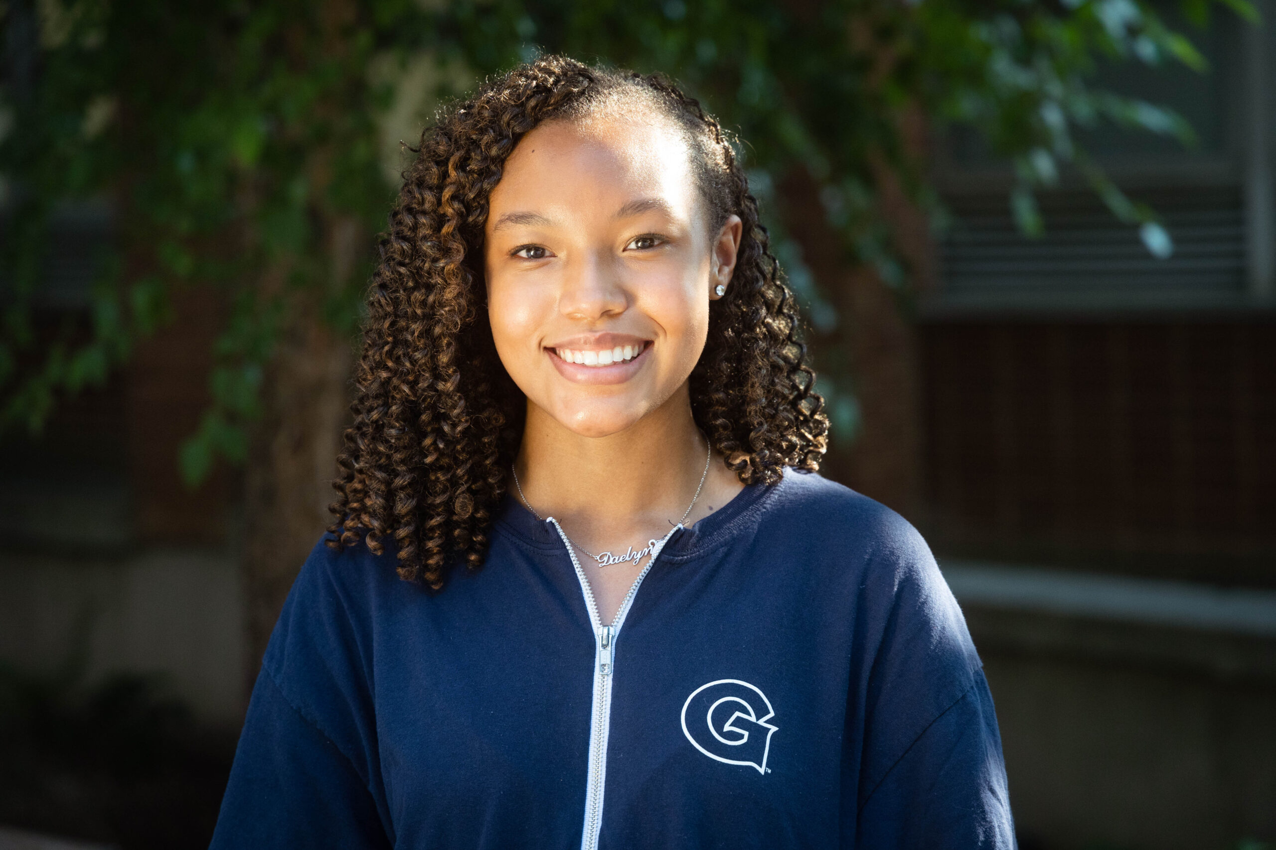 Headshot of Daelyn, wearing a GU athletics zip up smiling and standing in front of trees