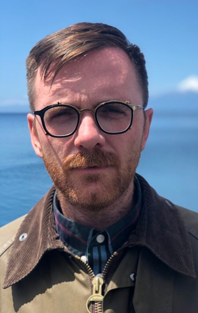 A bespectacled man stands in front of the ocean.