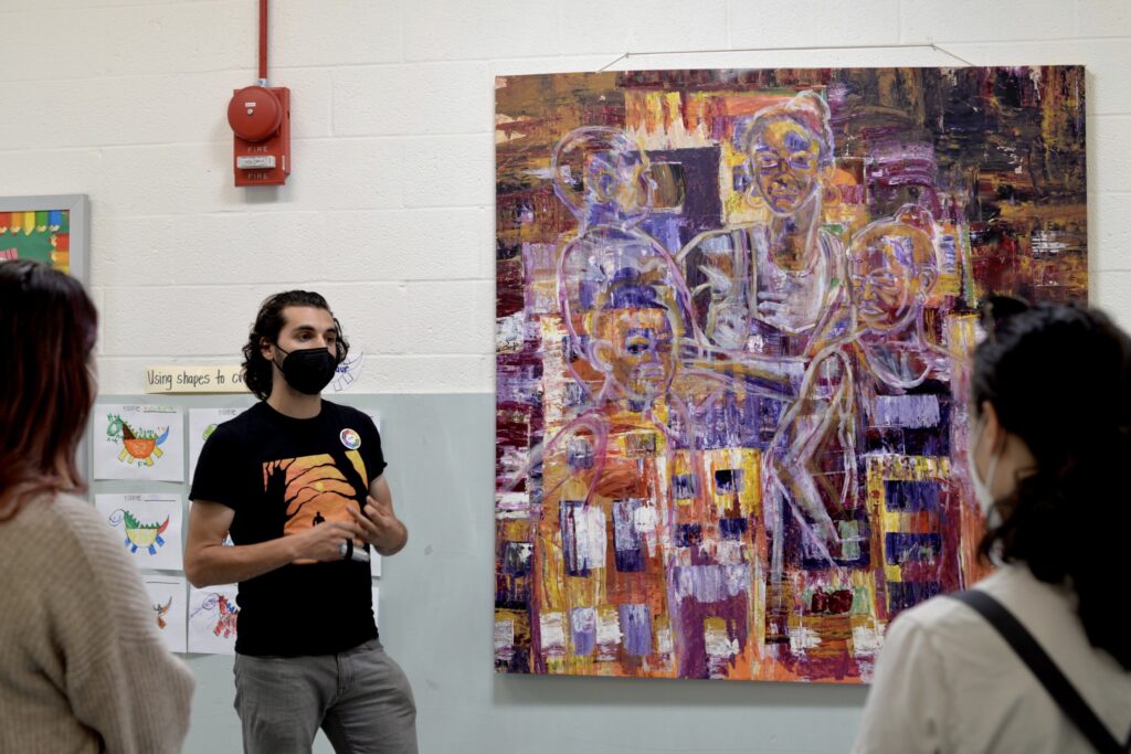 A man stands next to a painting and addresses a group of students. 