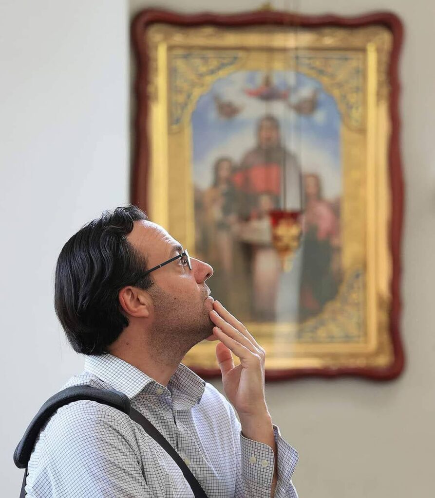 A man stands with his hand on chin, in the background is an out-of-focus religious painting.