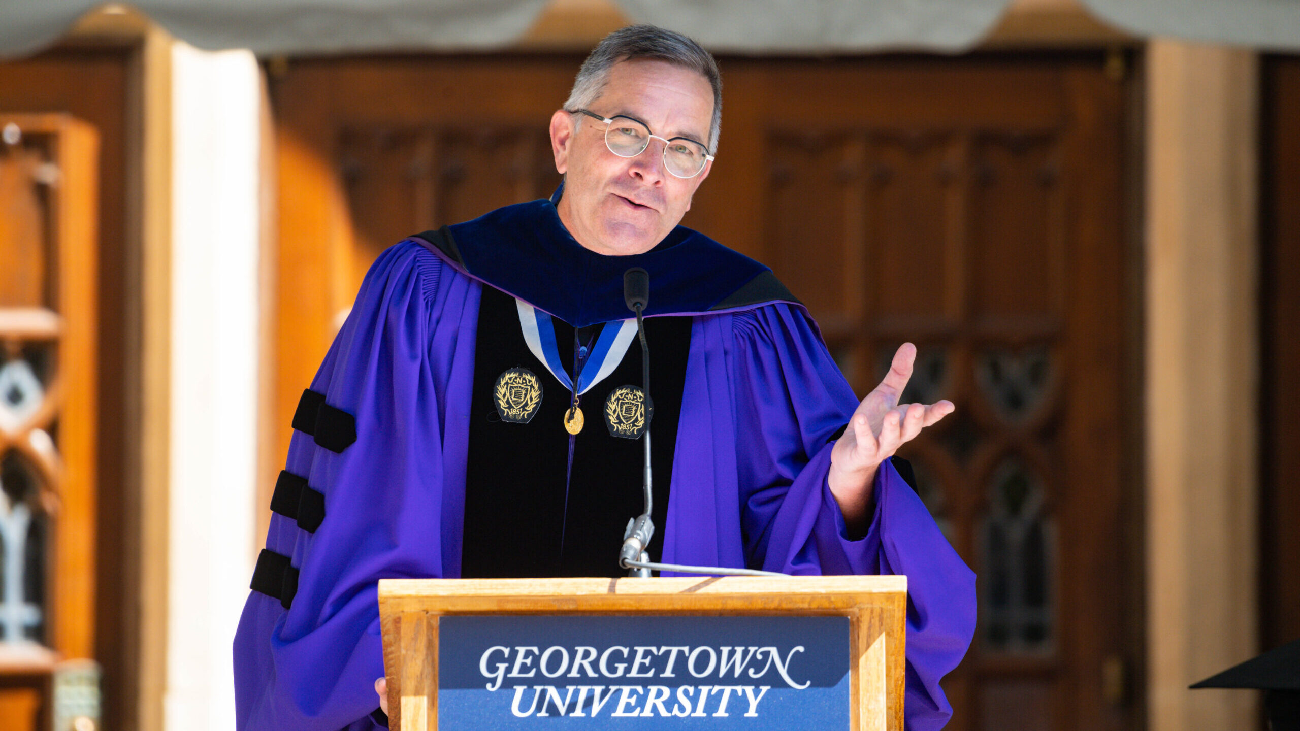 A bespectacled man in academic regalia speaks at a podium marked Georgetown University.