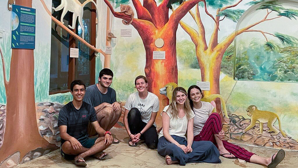 A group of students sits smiling in front of a wall painted with wildlife and covered with educational placards.