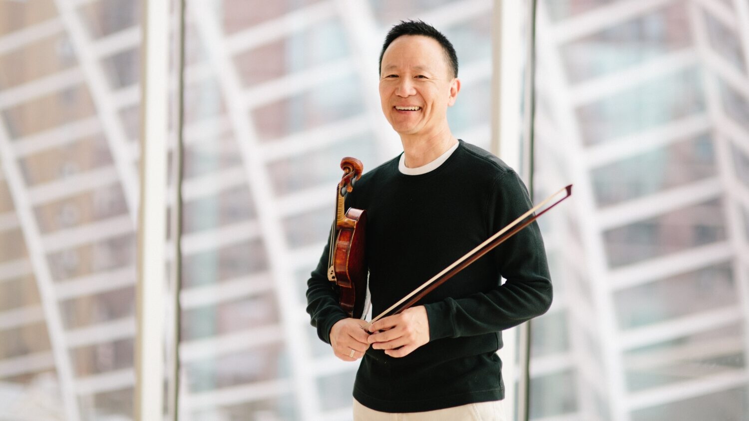 A man in a black sweater holds a violin and smiles