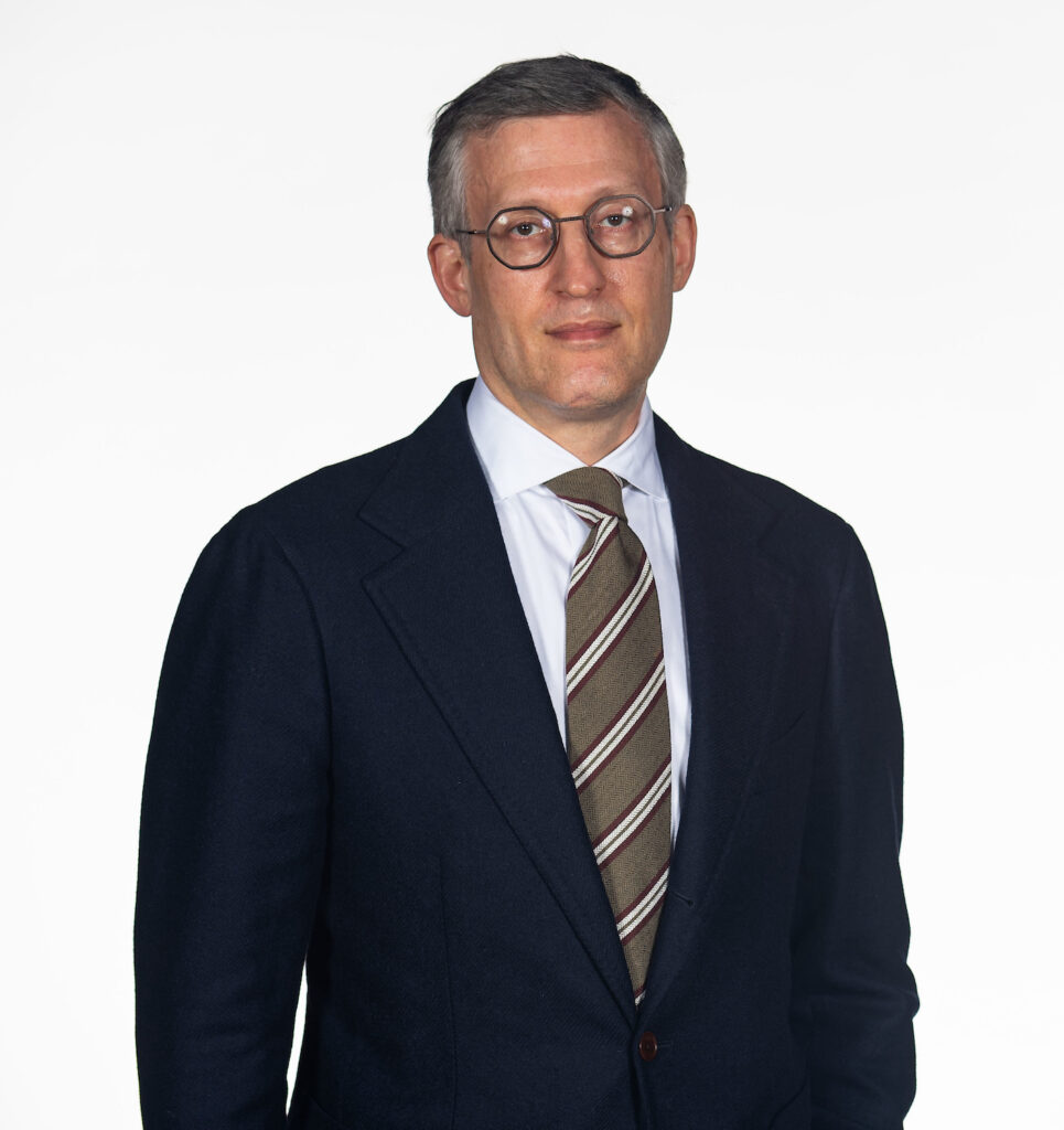 David Edelstein stands in front of a white background. He wears a blue suit jacket over a white shirt and striped tie. 