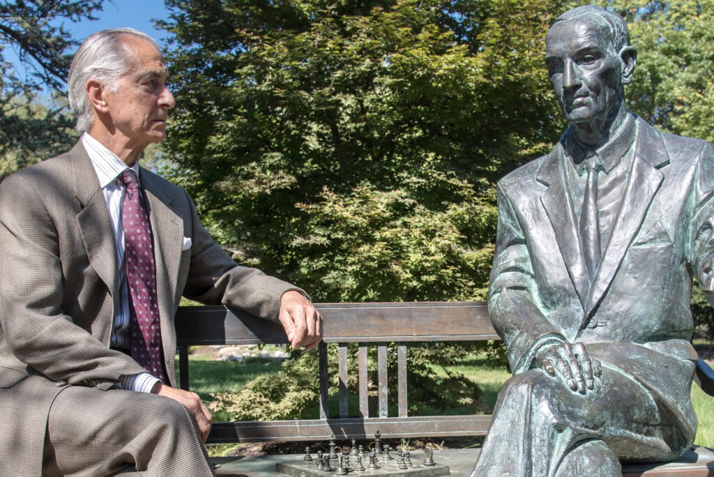 David Strathairn sits on a bench opposite the statue of Jan Karski. Strathairn wears a brown suit, striped shirt and red tie. 