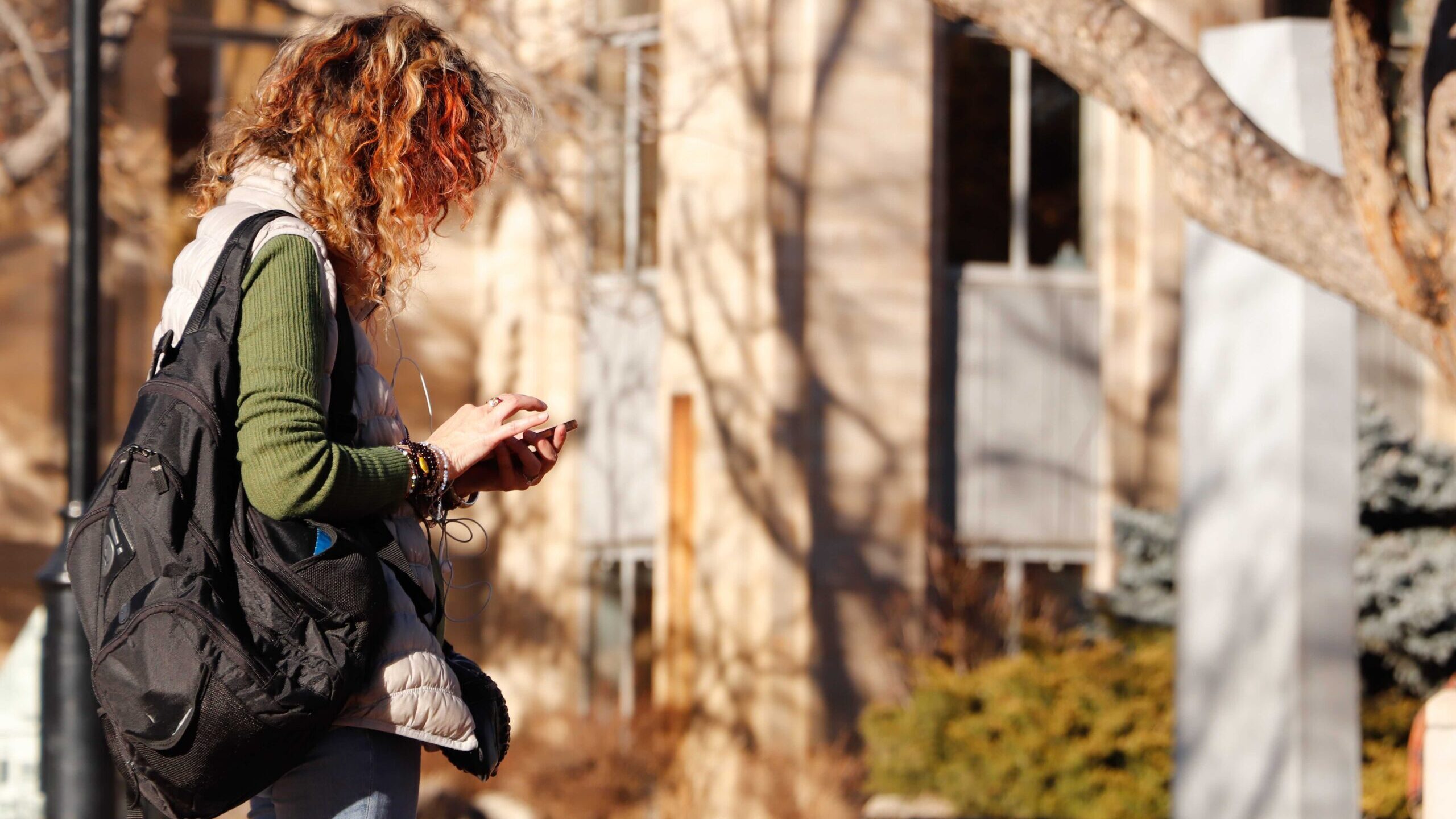 A woman with curly hair uses her smartphone. She stands in front of a blurry building and has a backpack slung over one shoulder.
