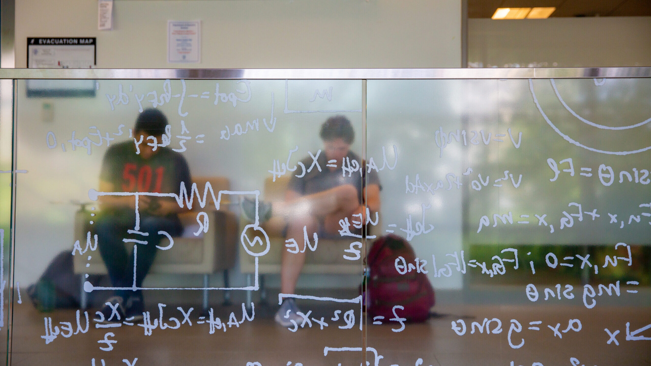 A frosted glass with equations on it obscures two blurred students studying in separate chairs.