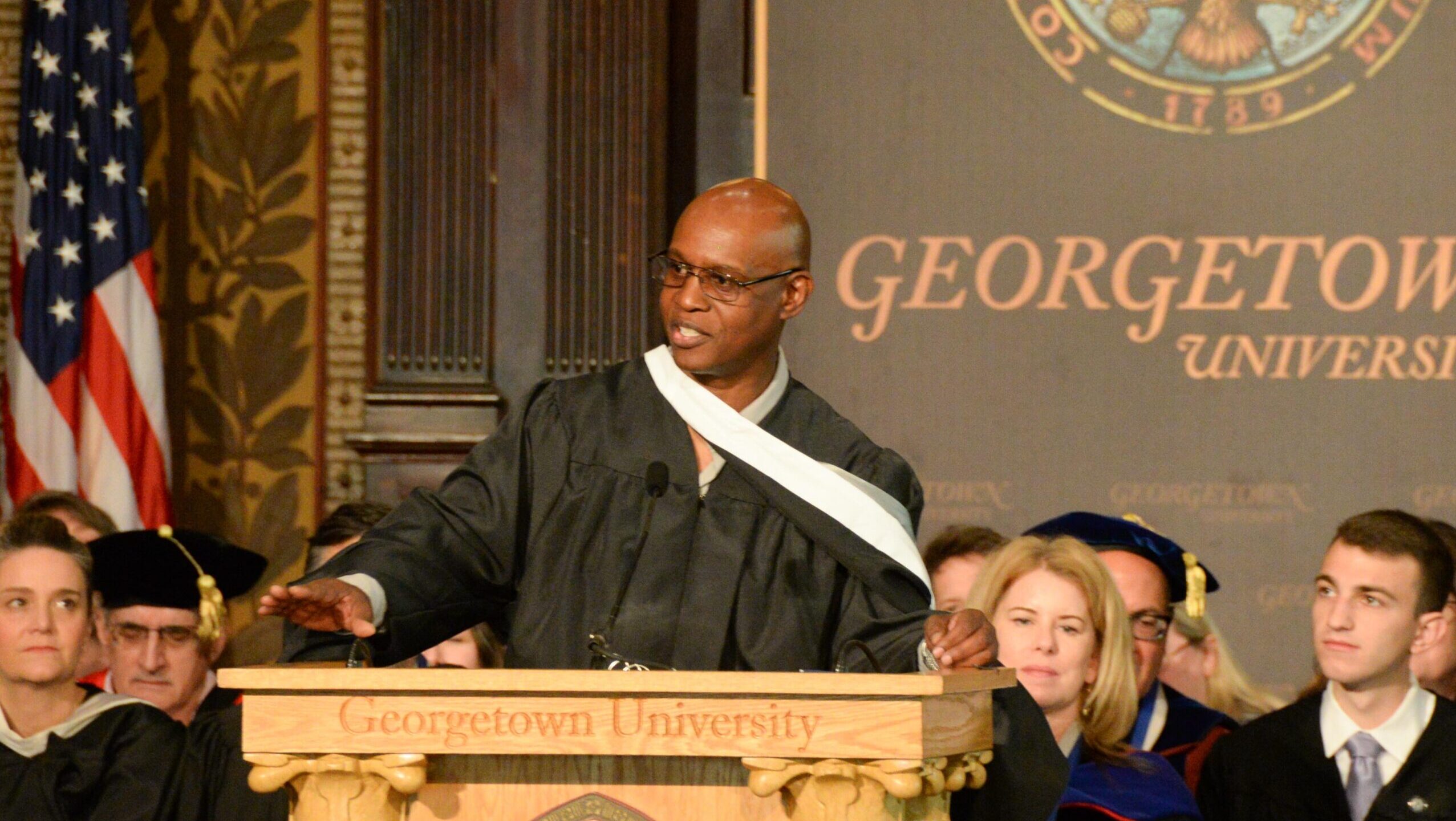A bespectacled man in academic regalia speaks at a podium.