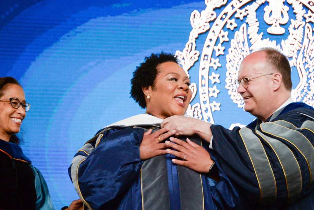 A woman with short dark hair is presented with an academic hood for an honorary degree. 