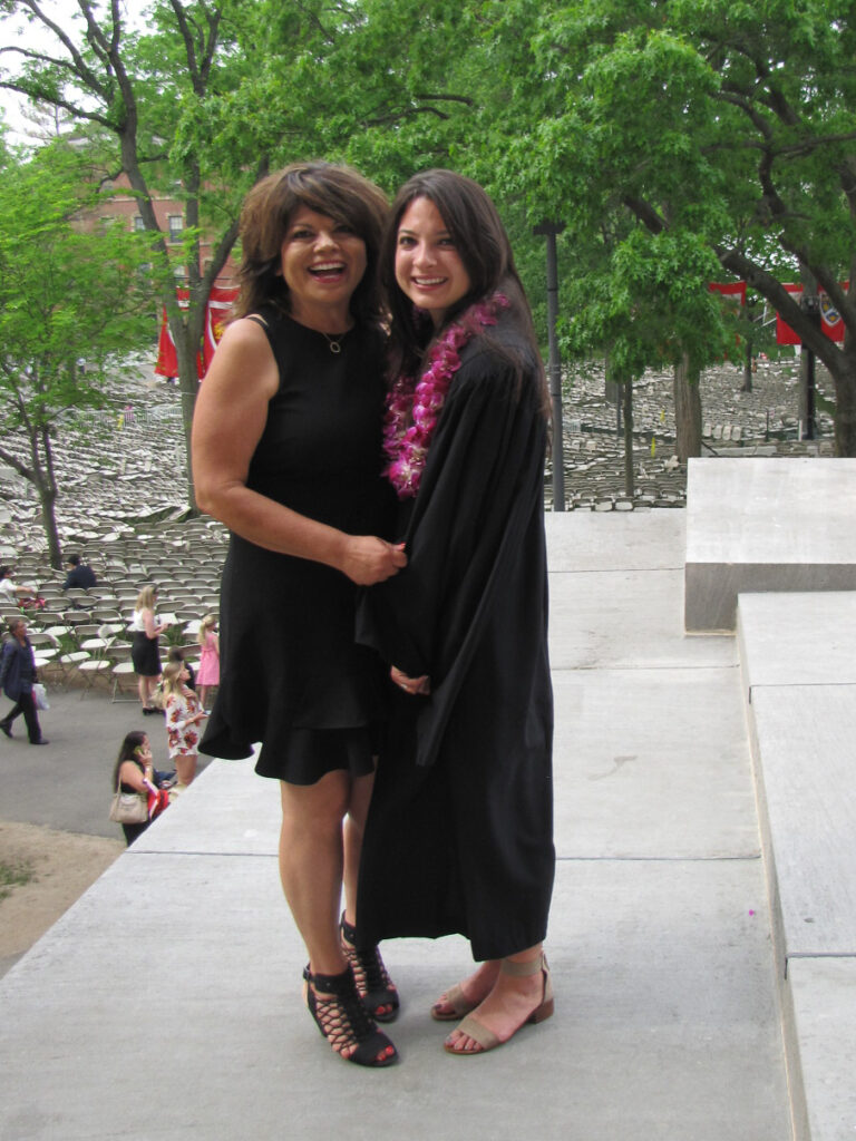 Two women stand smiling. One wears a black dress and the other is in academic regalia. 