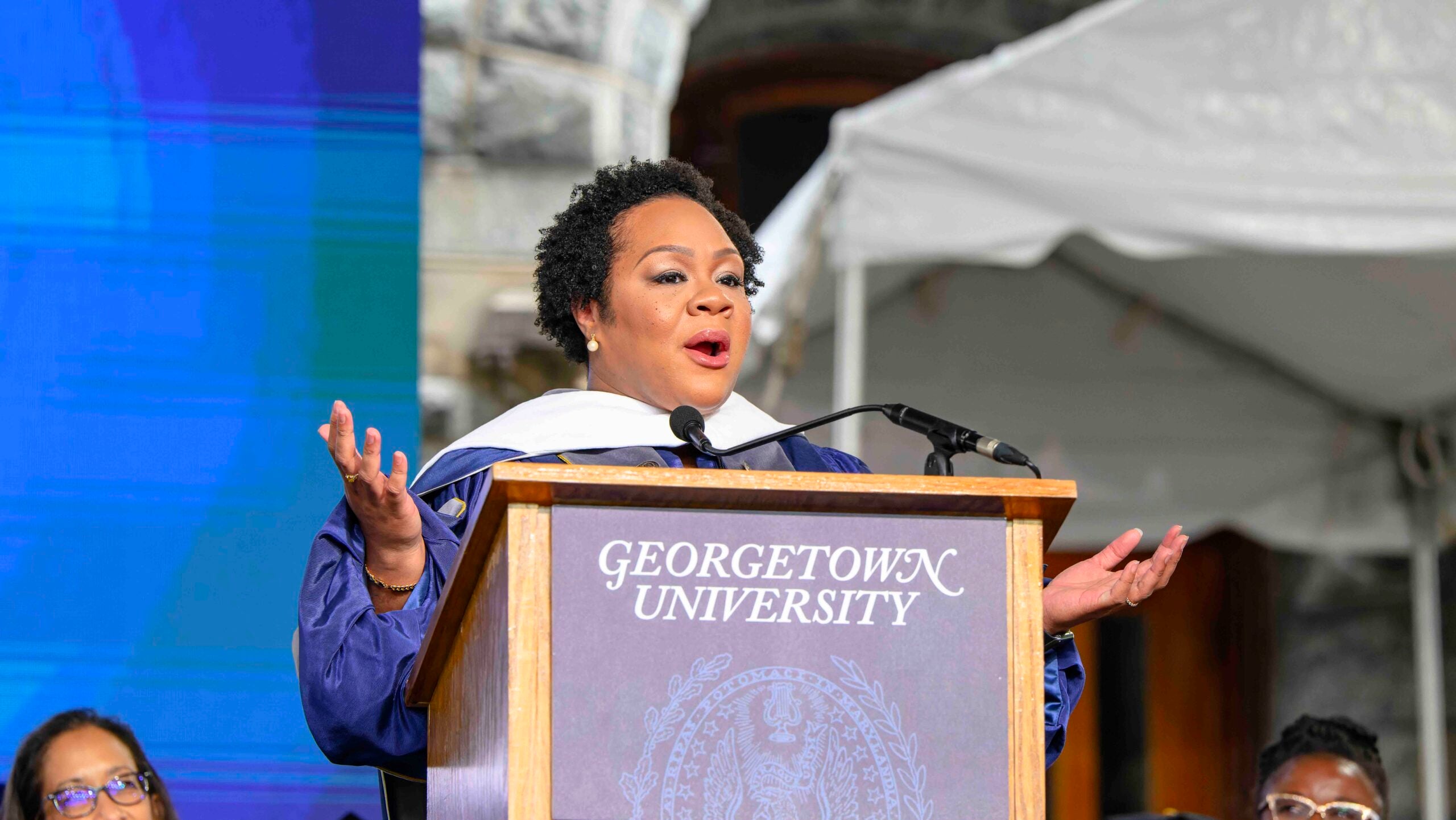 A woman with short, dark hair speaks at a wooden podium. She wears academic regalia.