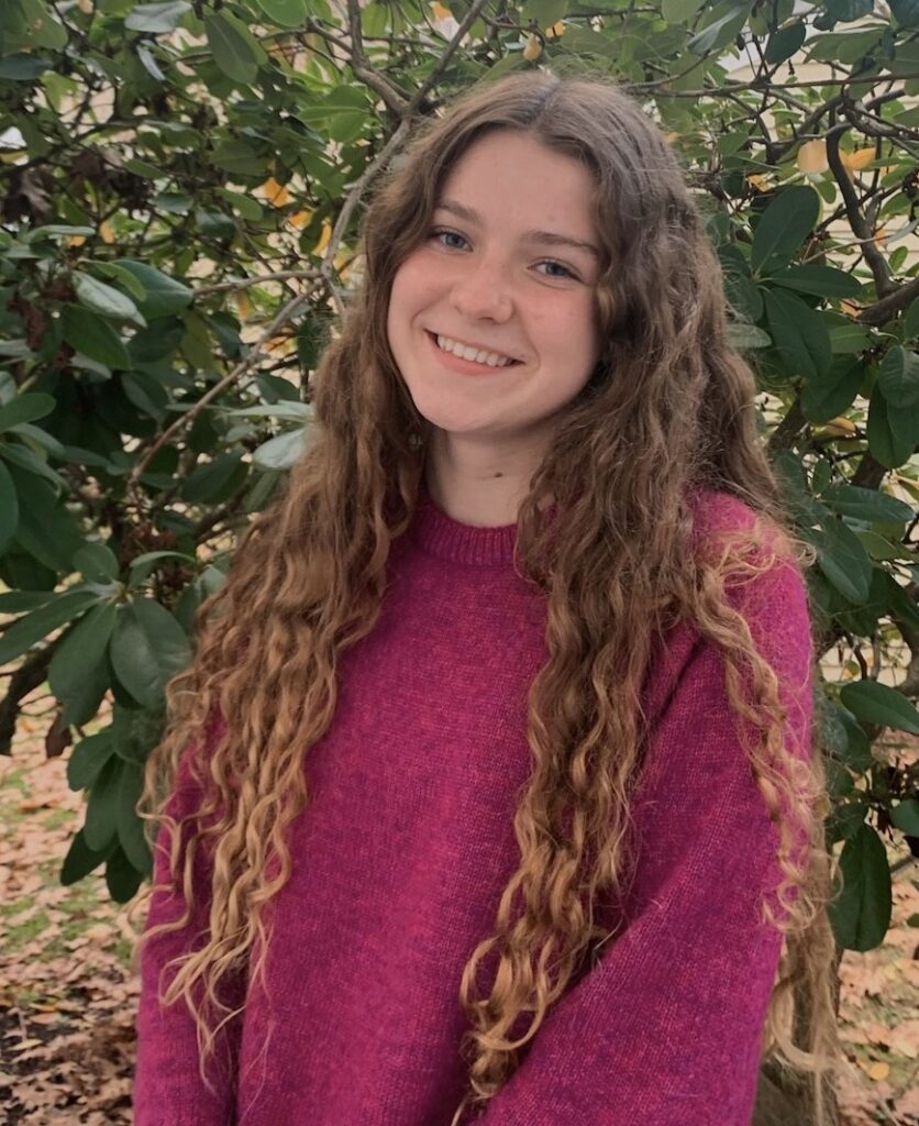 A girl with curly auburn hair smiles. She wears a pink sweater and stands in front of a green background. 