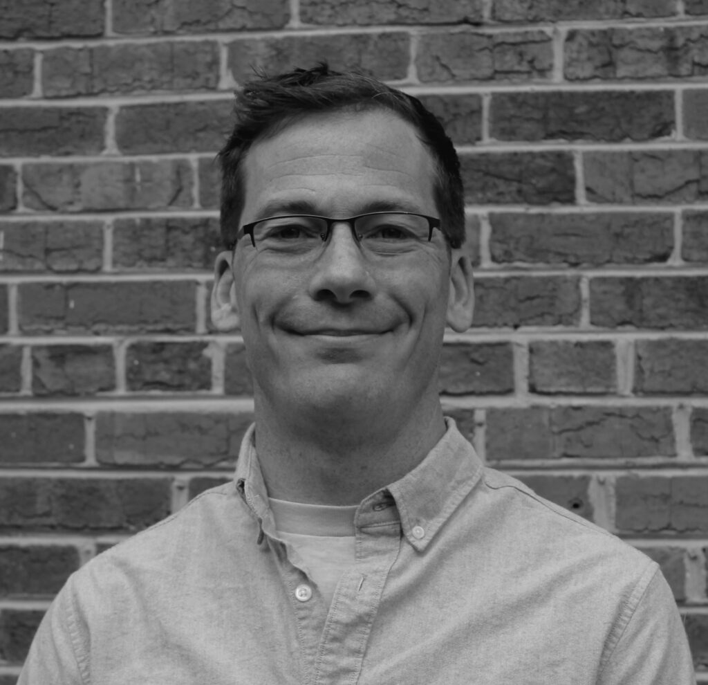 A bespectacled man wears a button-down shirt and stands in front of a brick wall. The photo is in black and white. 