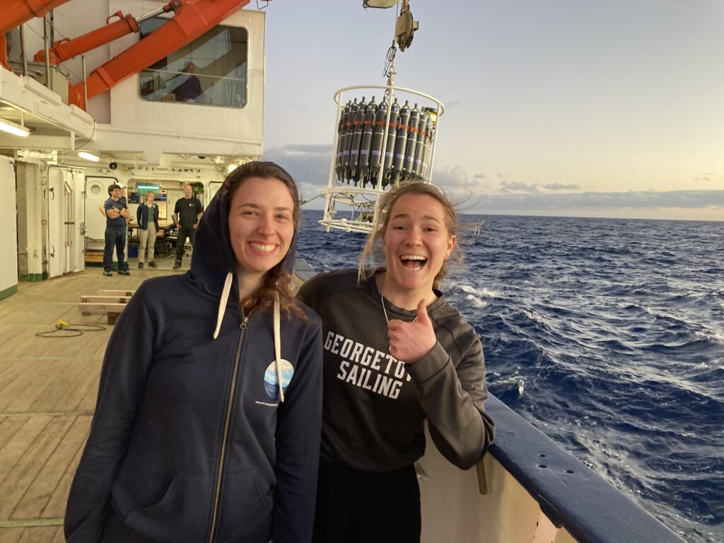 Two girls stand on the dock of a boat. One wears a Georgetown Sailing sweatshirt and is giving a thumbs up. Both are smiling. 