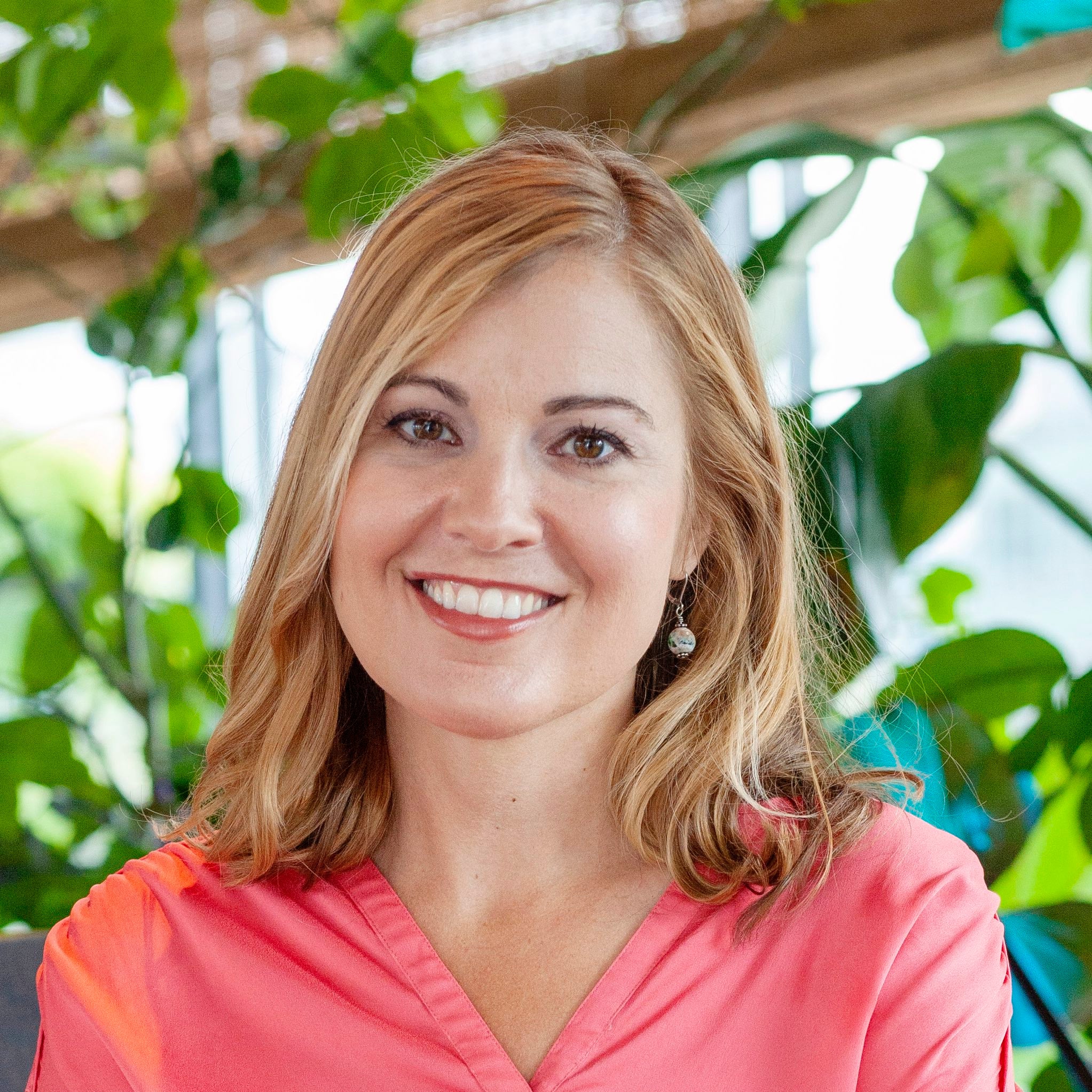 A woman with medium-length blonde hair smiles at the camera. She wears a pink blouse.