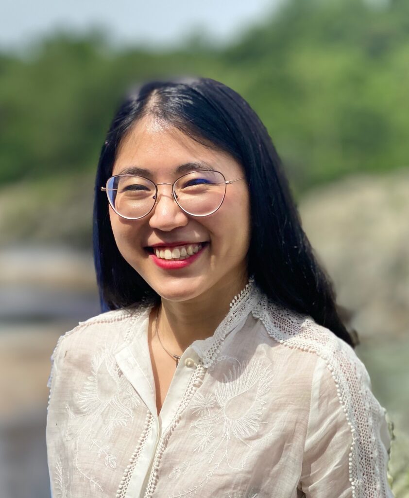 A bespectacled woman with long, black hair smiles. She wears red lipstick and a white blouse. 