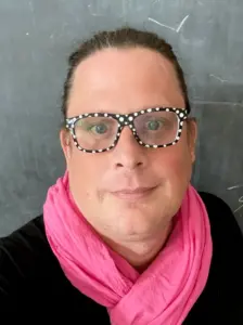 J softly smiles in front of a backboard, wearing a black top and pink scarf. J wears black-and-white polka-dot glasses. 
