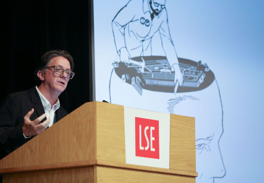 A bespectacled man speaks at a podium emblazoned with the letters LSE. He wears a white button-down shirt and a dark jacket. 