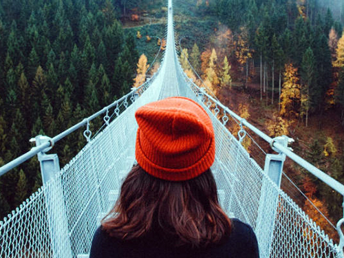 person in a knit orange-red cap with shoulder length brown hair peer out over a suspension bridge with trees visible on either side