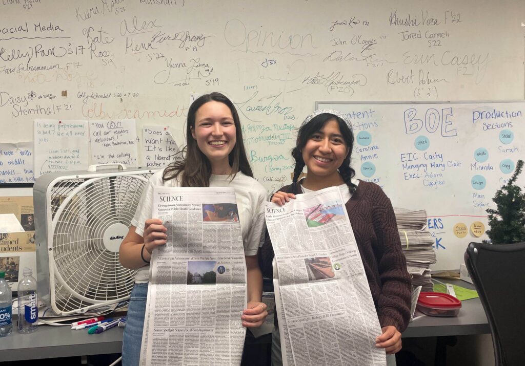 Two young women smile and hold up newspapers. They stand in front of a white board full of writing.