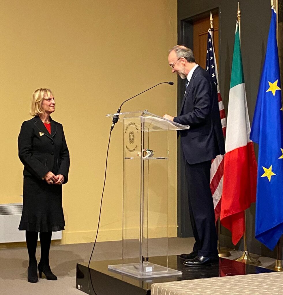 A woman in business attire stands as a man at a glass podium speaks. Behind the man there are three flags: The American Flag, the Italian Flag, and the European Union Flag.