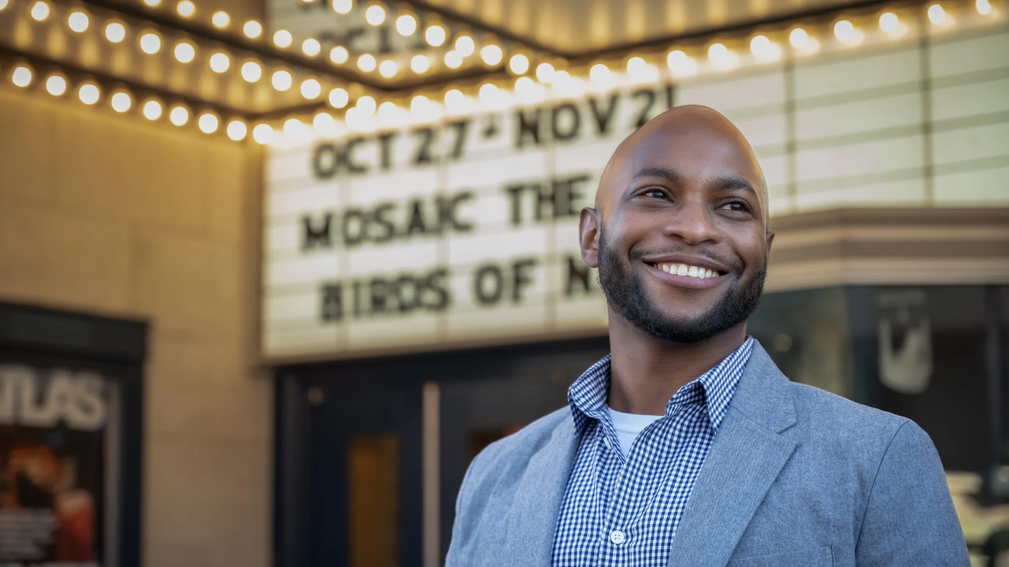 A man with a well-groomed beard smiles in front of a theater marquee.