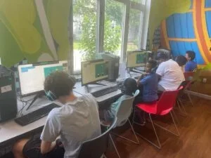 A line of students sit at desktop computers. The students are in a computer lab where the walls are painted green. 