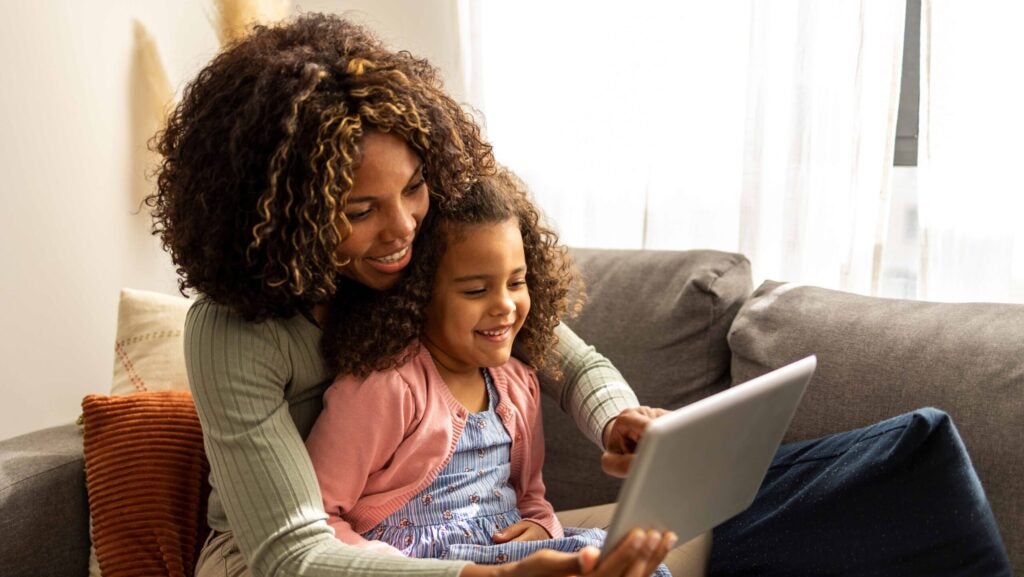 A mother and daughter sit together on a couch. The daughter sits in the mother's lap and both collaboratively look at a tablet.