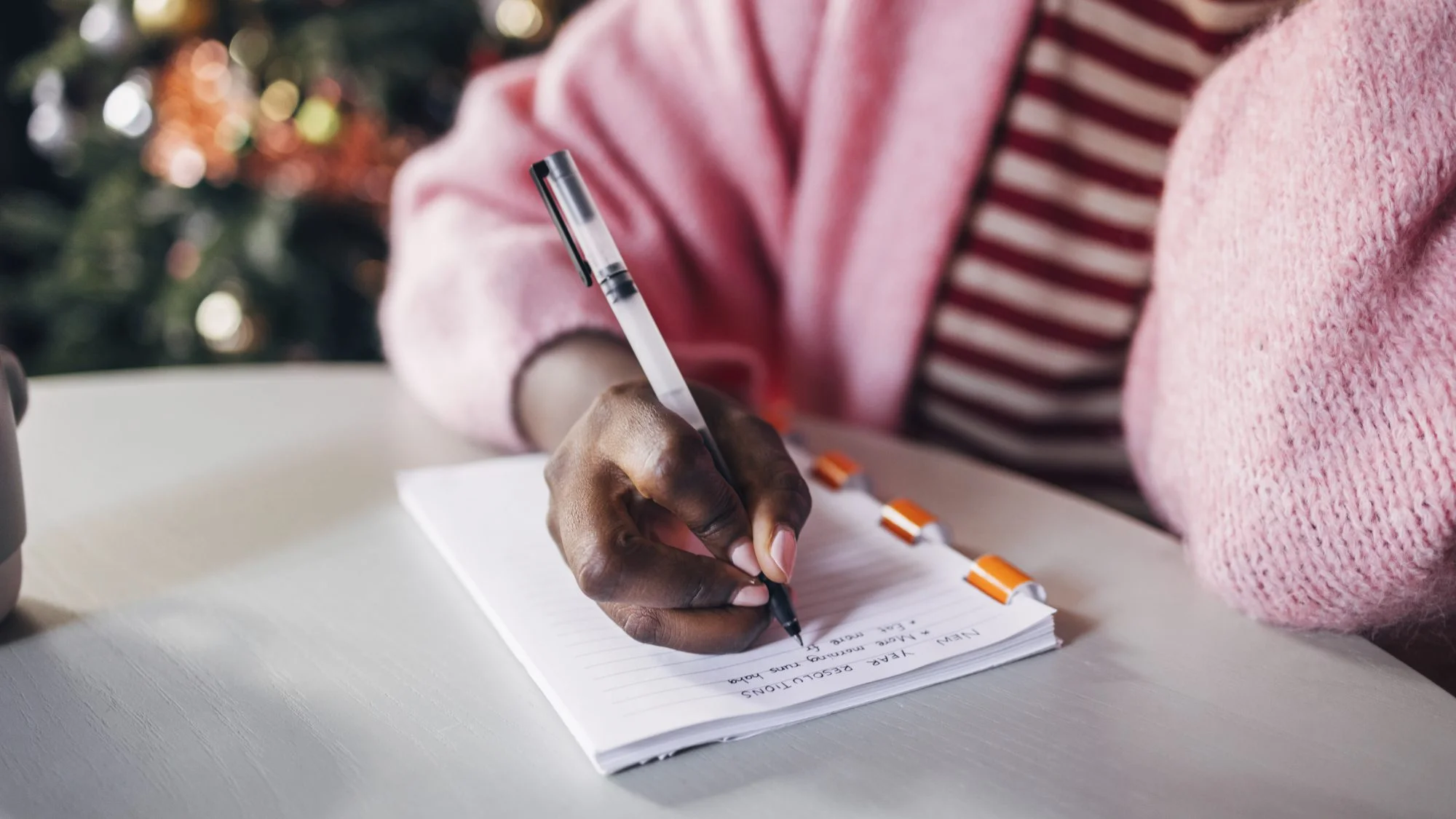 A Black woman wearing a pink sweater and striped pink shirt writes her New Year's resolutions on a notebook.