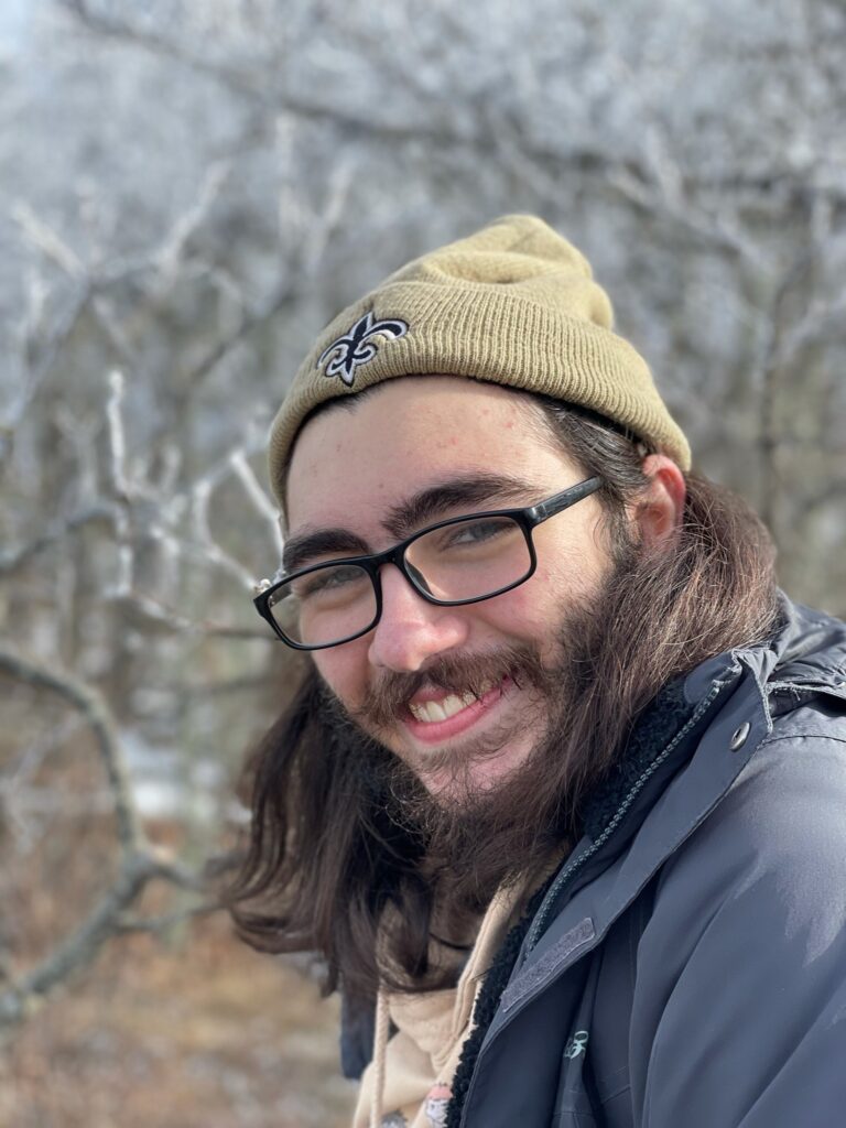 A bespectacled man with long, dark hair looks over his shoulder and smiles at the camera. He stands outside -- there are out-of-focus trees behind him. He wears a knit cap emblazoned with the fleur-de-lis and a navy blue windbreaker. 