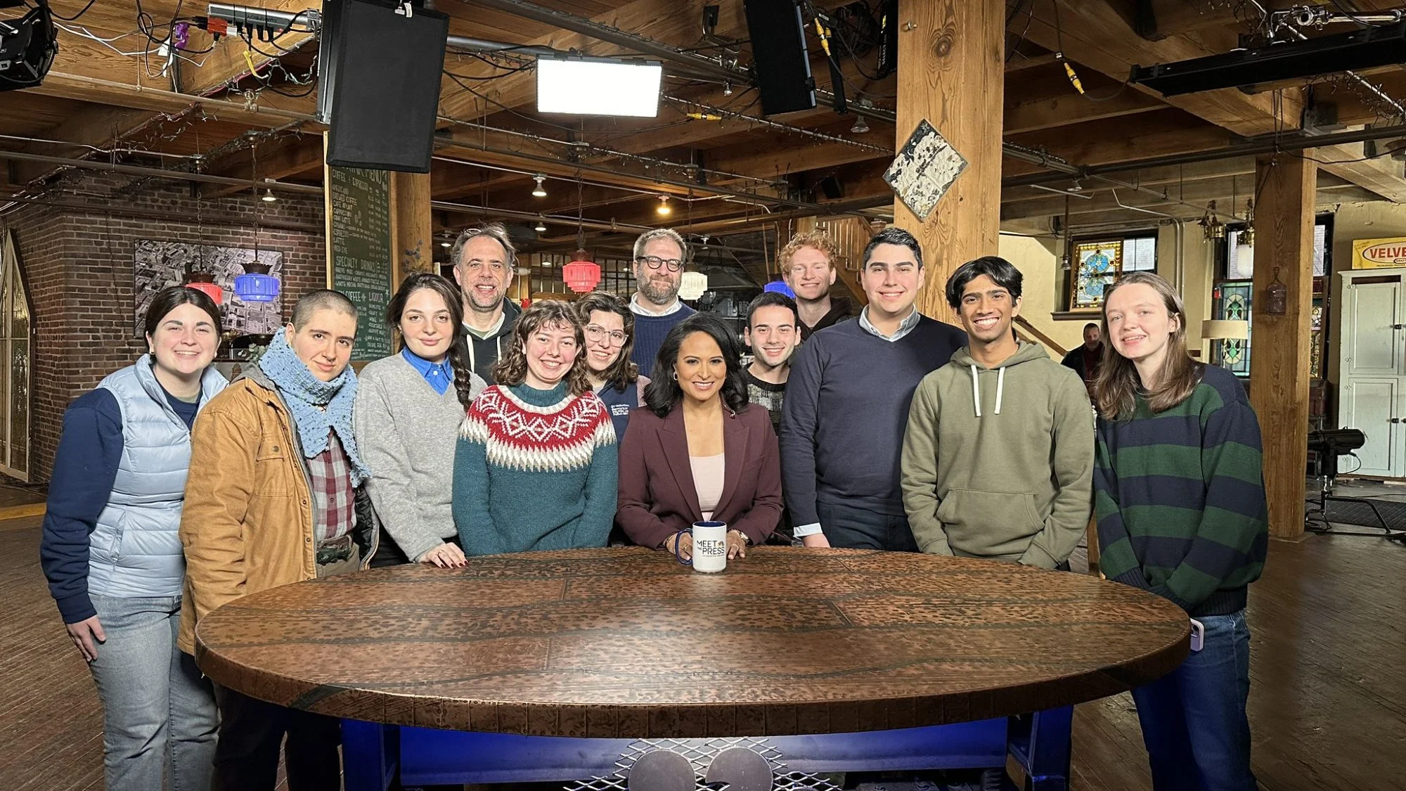 A group of students stands around a wooden desk with the NBC logo. There is camera equipment around the desk signifying that this is a television set.