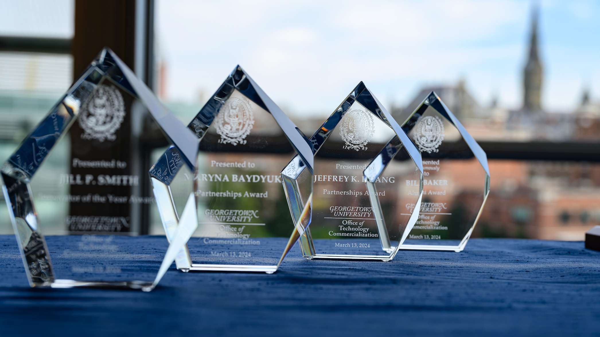 Glass awards arranged on a blue tablecloth. The out-of-focus outline of Healy Hall is in the background.