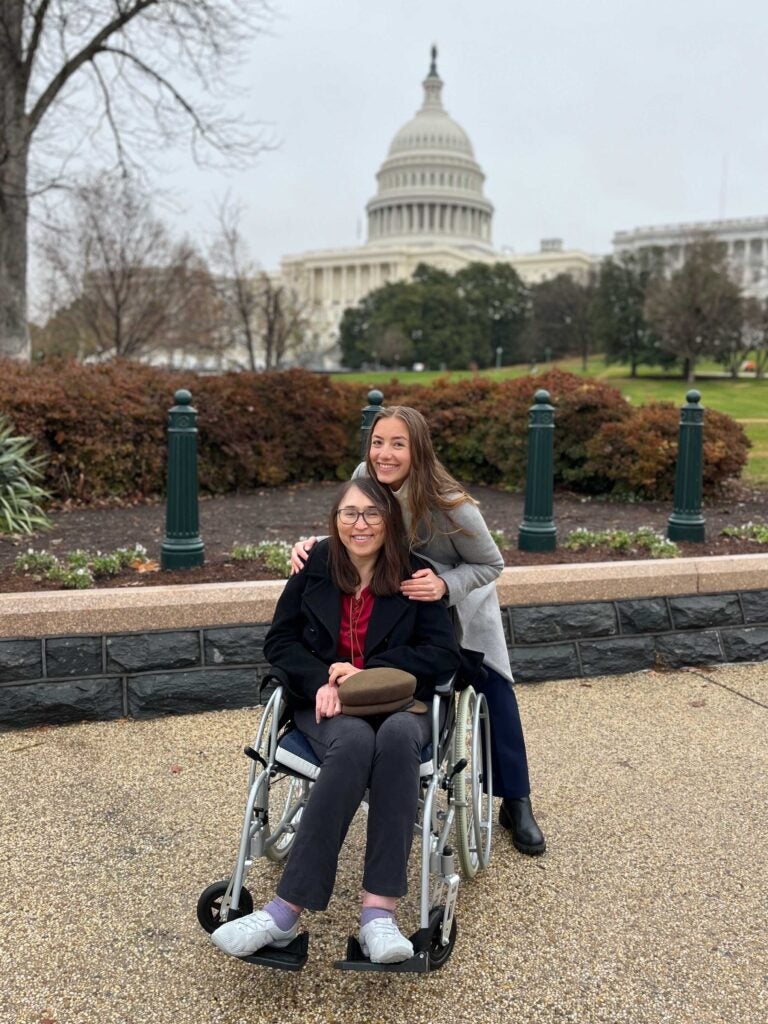 A woman with long, auburn har stands behind a woman in a wheelchair with long, dark hair. They both are smiling and looking at the camera. The U.S. Capitol is behind them.