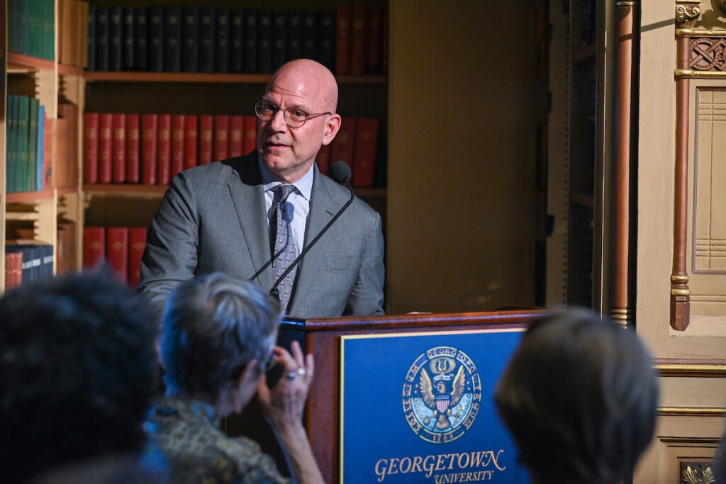 A man in a gray stand speaks at a podium emblazoned with the Georgetown University Seal. He wears glasses and stands in front of an elegant library.