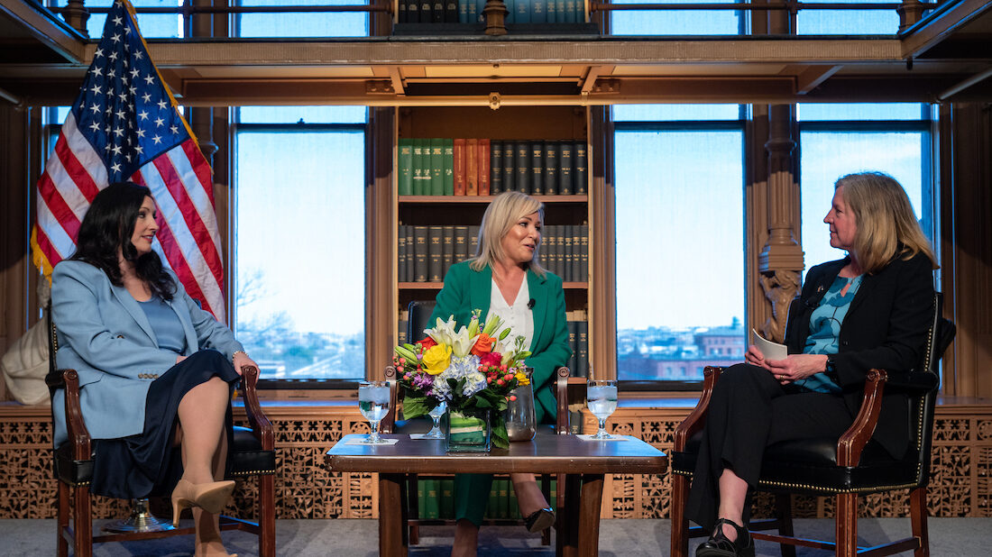 Three women in formal business attire sit in an elegant library. In the middle there is a table with flowers. The American flag is in the background.