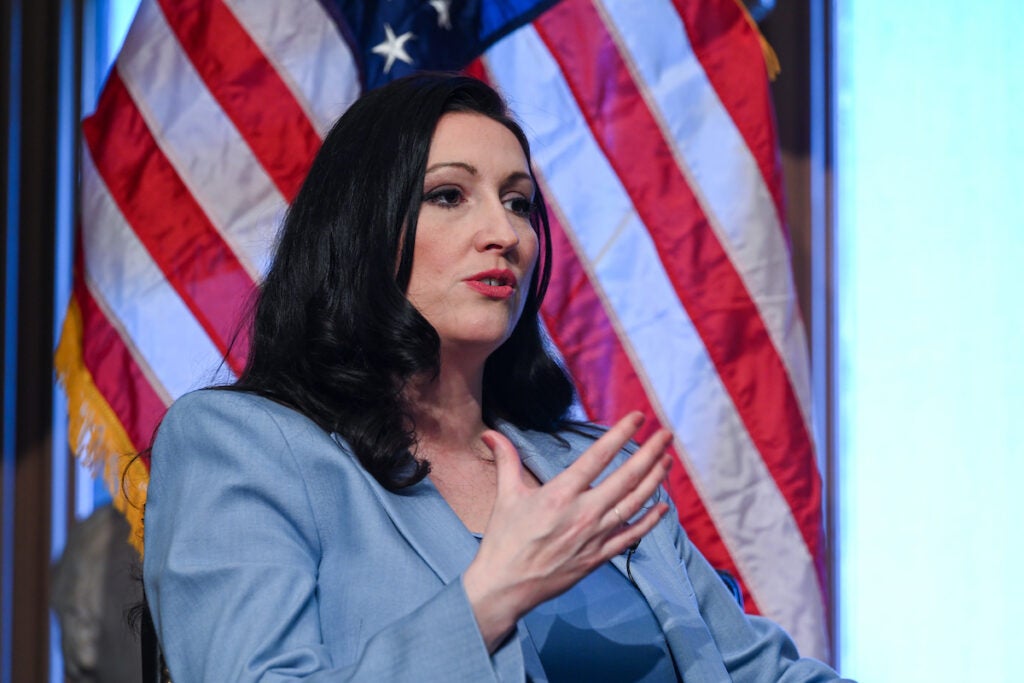 A woman with long, dark hair speaks in front of an American flag. She gesticulates with one hand and wears a baby blue suit jacket. 