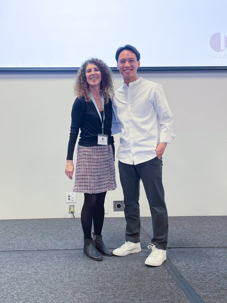 A woman and a man stand together and smile in a conference room. The woman wears a colorful skirt and a dark top. She has long, curly hair. The man wears a white button down shirt and dark pants. His hair is short and combed with a middle part. 