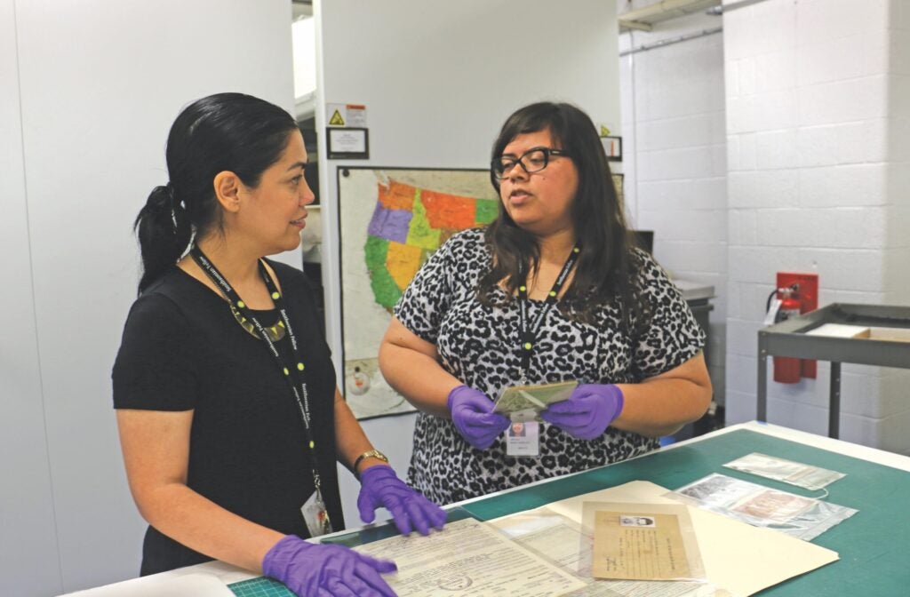 Two woman stand at a table examining old documents that are encased in plastic. Both women wear rubber gloves and their hair is back.