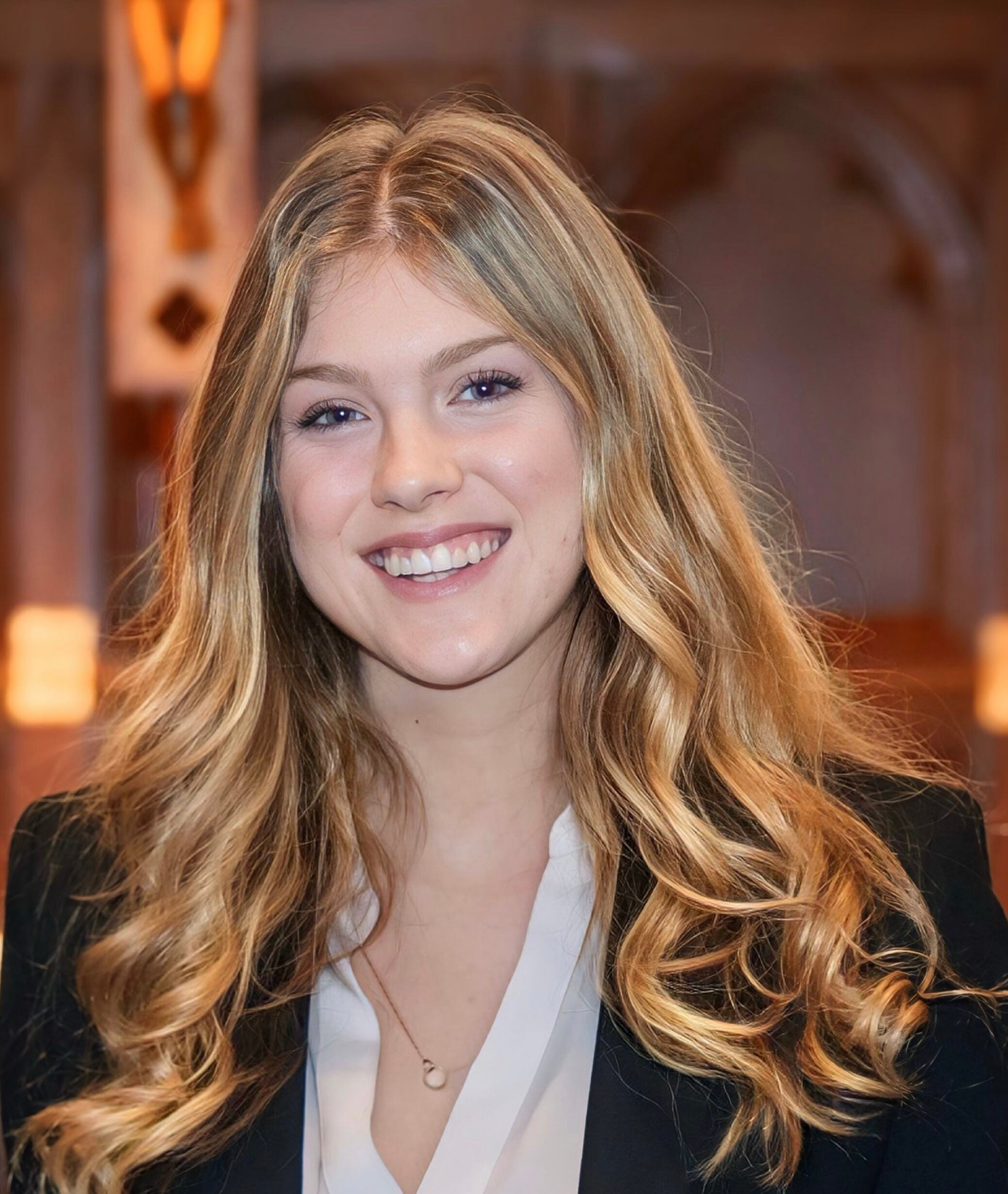 A girl with long, blonde hair smiles inside. The background is out-of-focus wood paneling. She wears a white blouse and a black, formal jacket. 