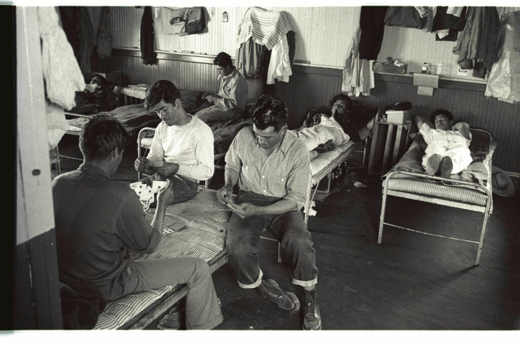 A black-and-white photo of several men in a sleeping quarters. Some rest on beds while others smoke cigarettes and play cards on a cot. 