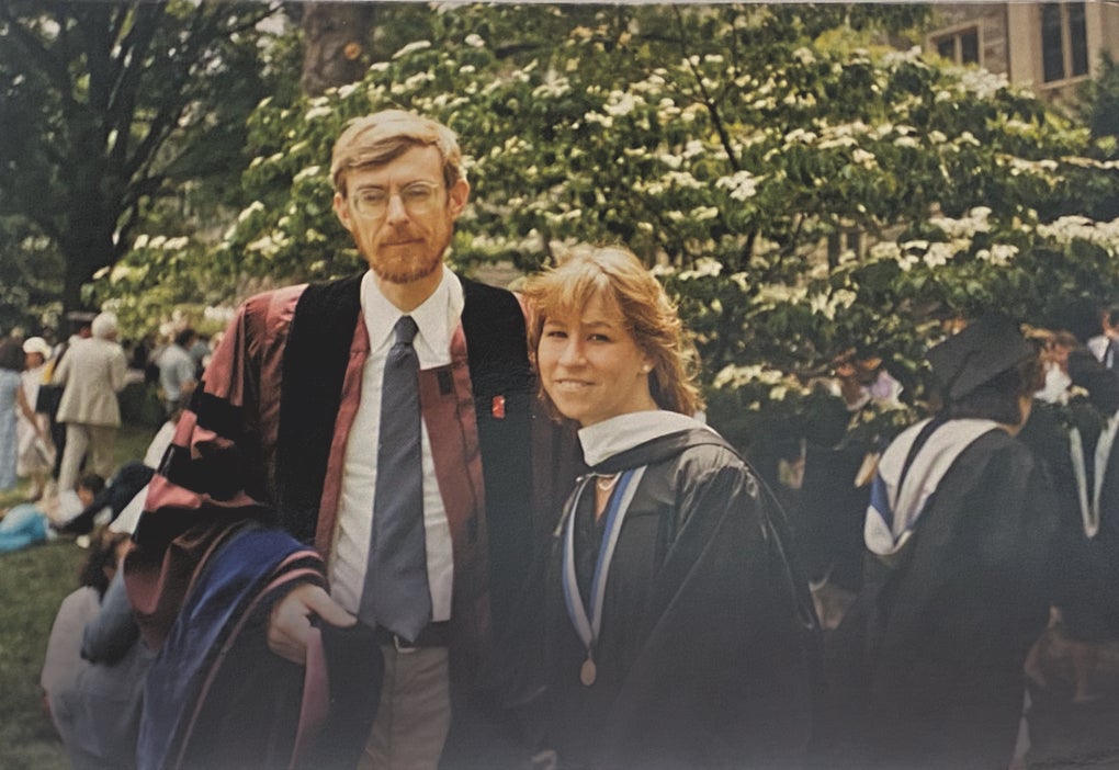 Two people pose in academic regalia. One, a man, is taller and wearing glasses. The other, a woman, has medium-length blonde hair. 