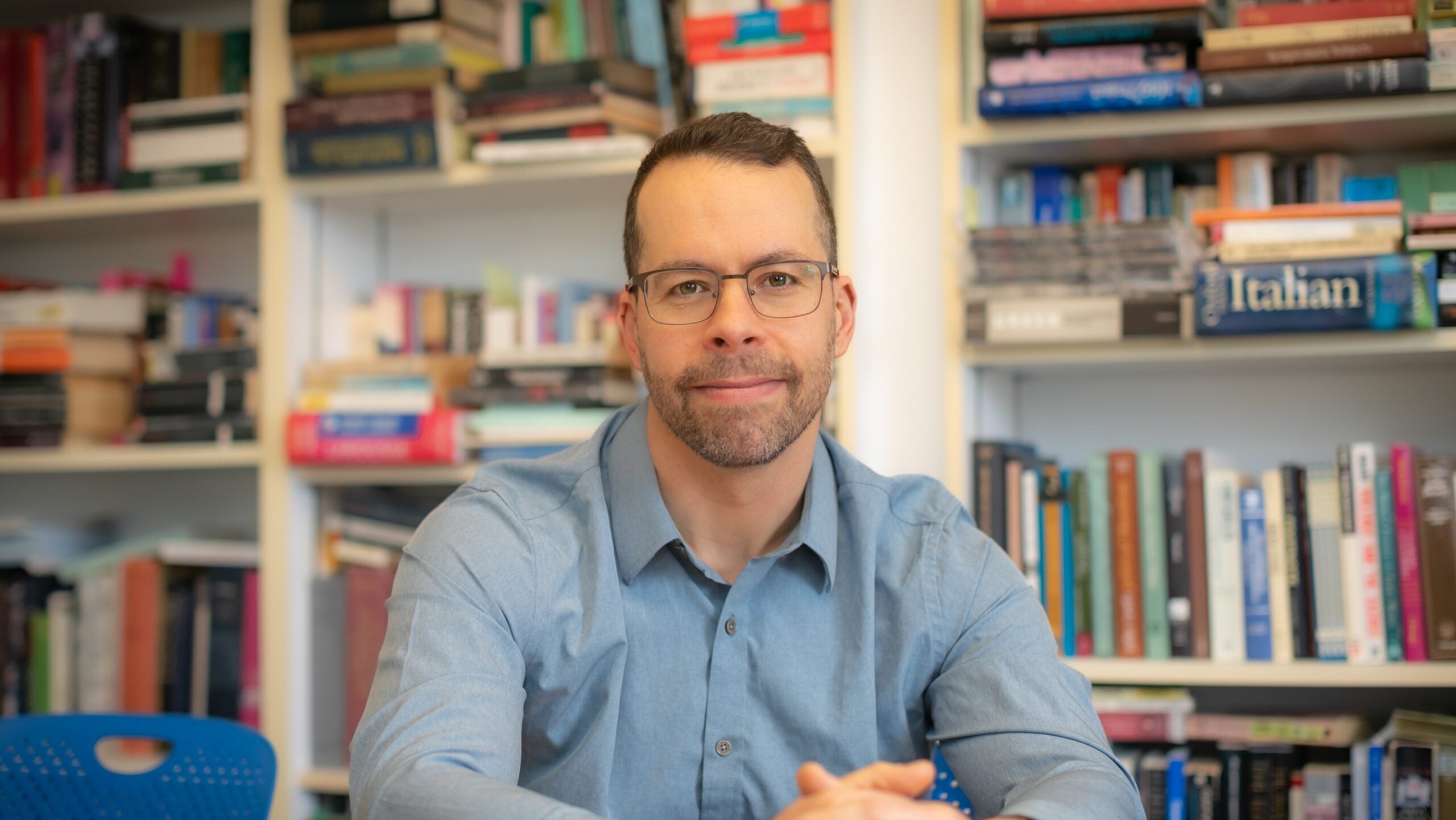 A bespectacled man softly smiles at his desk. He wears a light blue shirt and is surrounded by overflowing bookshelves.