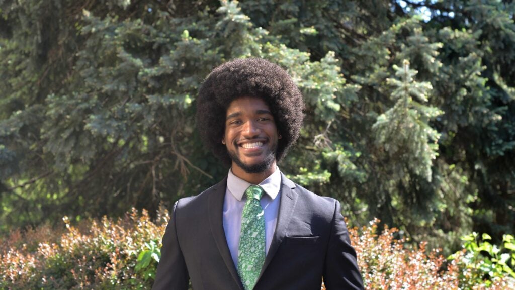 A man wearing a black suit and green tie smiles outside. The tie is patterend with different kinds of leaves. He sports an afro and a short beard.