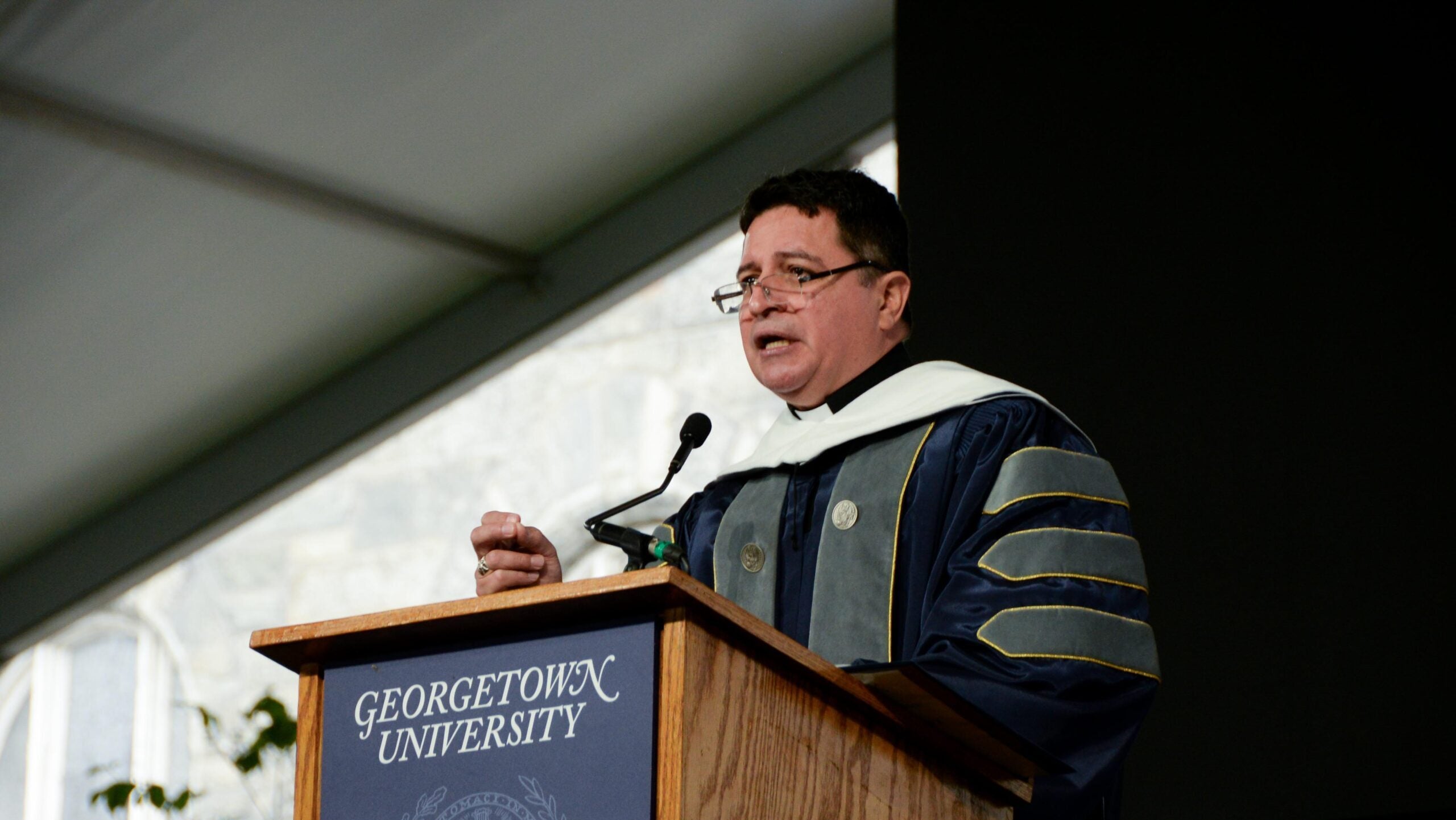 A bespectacled man in blue and gray academic regalia speaks at a lectern.