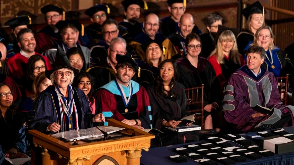 A crowd of faculty in academic regalia on a stage. A woman speaks at a podium and dons a witch hat.